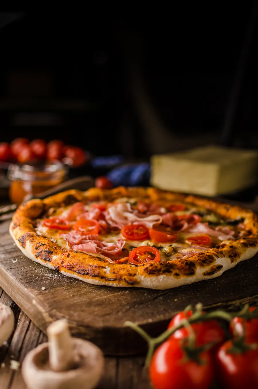 Pizza sa pršutom i čeri paradajzom na drvenoj dasci, u pozadini sir i paradajz