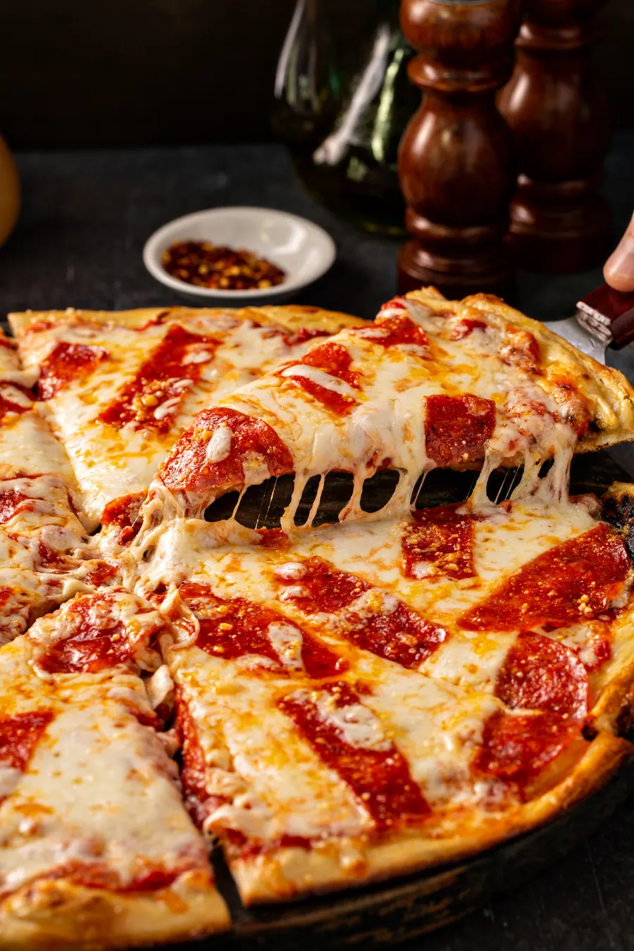 Parče pizze sa rastegnutim sirom podignuto sa tacne, u pozadini ljute papričice i mlin za biber